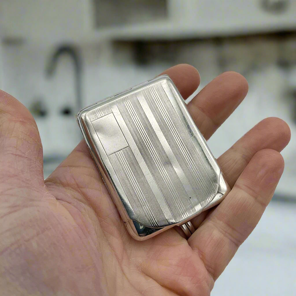 Silver matchbook case with a lined pattern and blank box for engraving initials in left hand corner held in a hand.