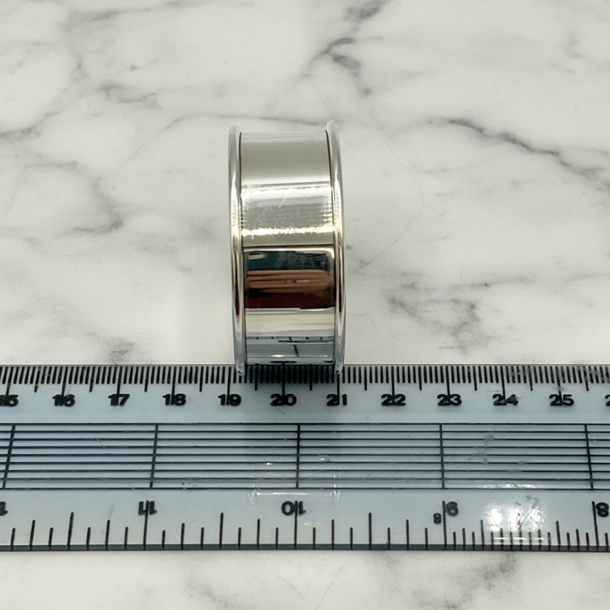 A polished silver napkin ring standing upright on a white surface, positioned above a clear plastic ruler to show its scale.
