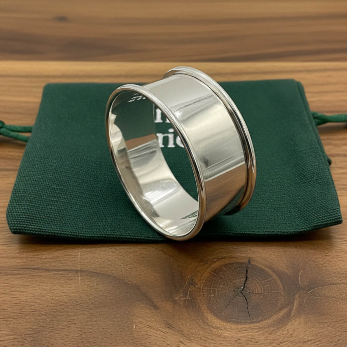 A polished silver napkin ring resting on a dark green drawstring pouch against a plain wood background.