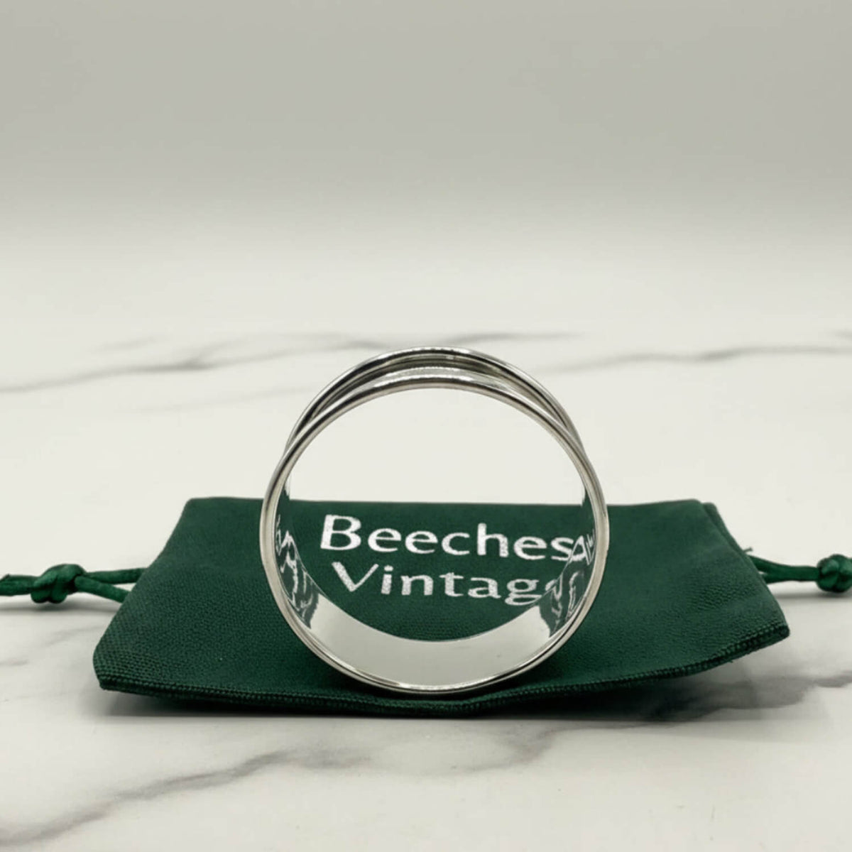 A polished silver napkin ring resting on a dark green drawstring pouch with the brand name 'Beeches Vintage' printed on it, set against a light gray marble background.