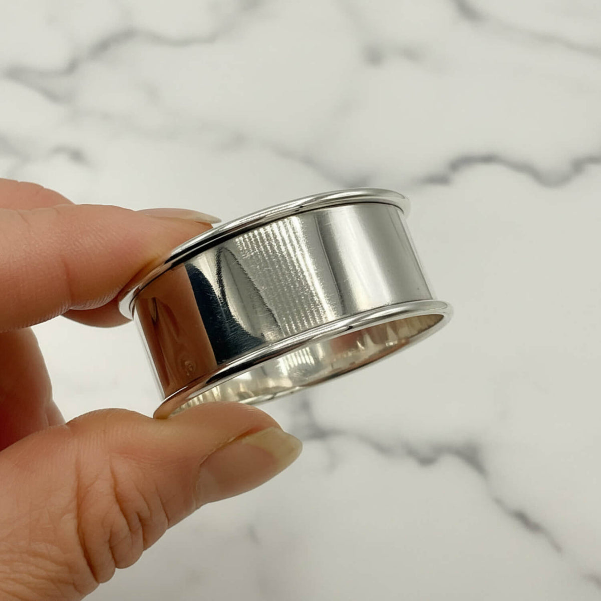 A polished silver napkin holder with a textured inner band, held between fingers against a plain light background.