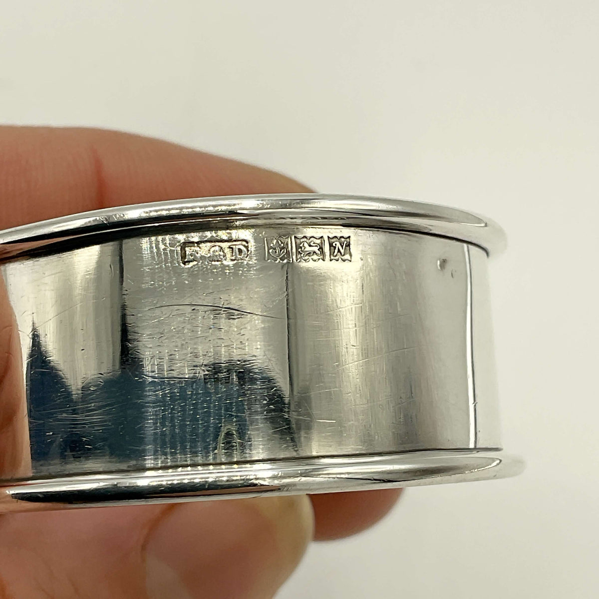 A close-up of a polished silver napkin ring held in a hand, showing engraved hallmarks on the side against a plain light background.