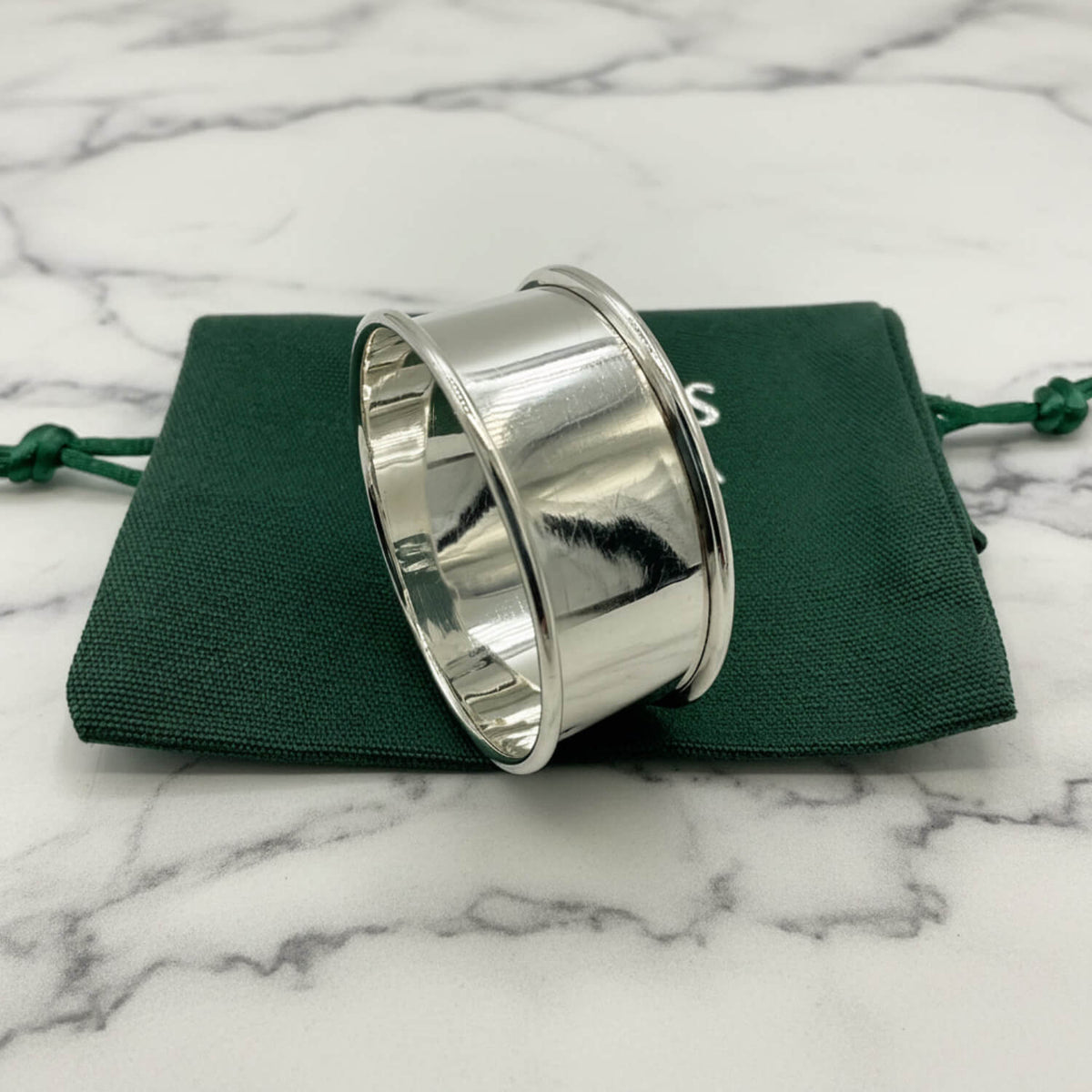 A polished silver napkin ring, resting on a dark green drawstring pouch against a light gray background.