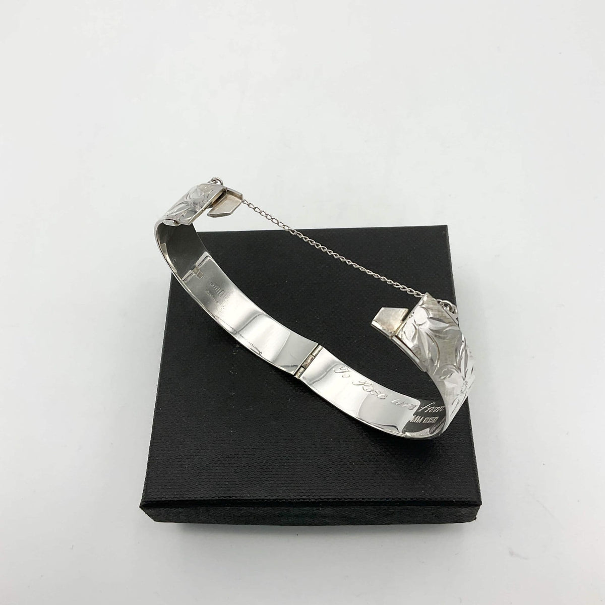Silver bracelet with engraved flowers with clasp open showing the safety chain sitting on a black box
