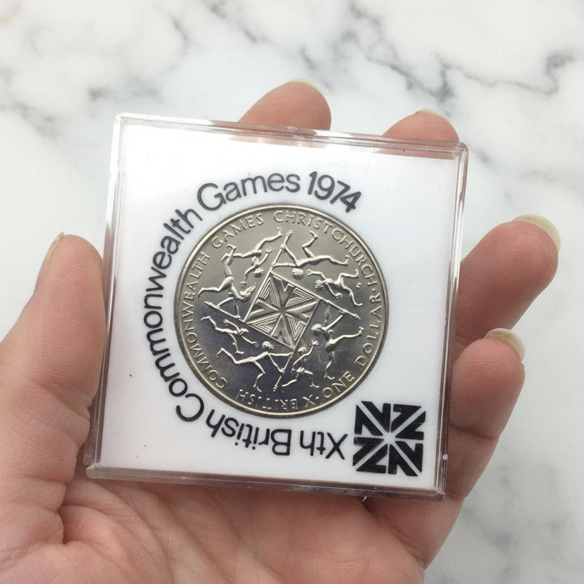 One dollar coin in a white case with 10th British Commonwealth Games 1974 around it held in a hand