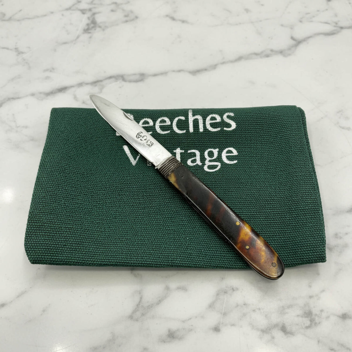Antique fruit knife with a silver blade and a dark brown faux tortoiseshell handle, resting diagonally on a green canvas pouch with white text that reads 'Beeches Vintage'.