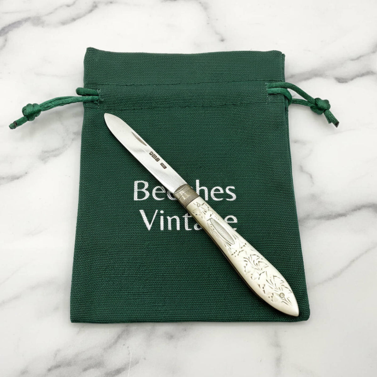 Decorative silver knife with ornate handle on a green drawstring bag labeled 'Beeches Vintage' on a marble surface.