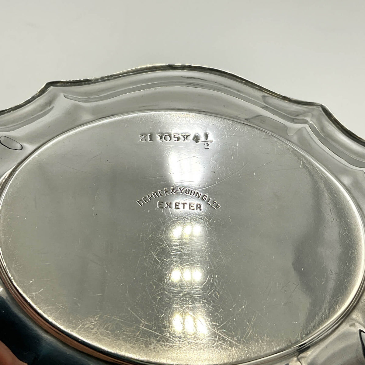 Close-up of the bottom of a silver metal dish or plate with a scalloped rim, showing engraved maker's marks and weight/strength text on a light gray background.