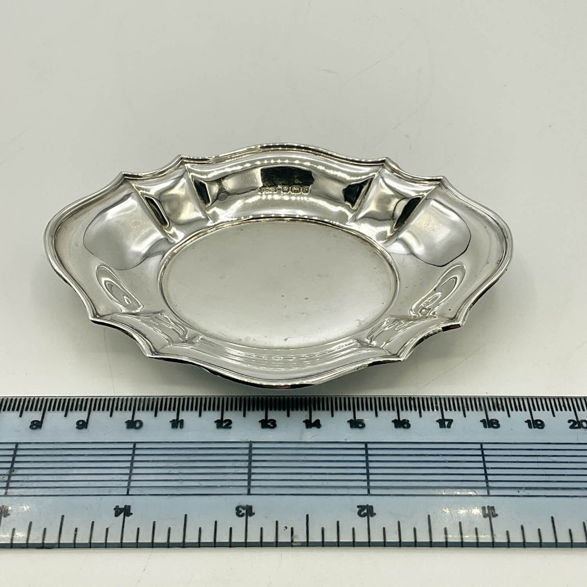 A small oval silver dish with a scalloped rim, shown on a white background with a ruler at the bottom for scale.