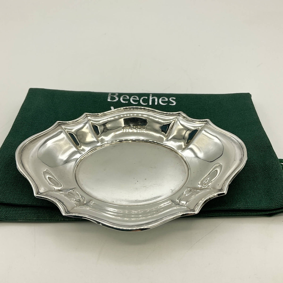 A polished silver oval serving tray with decorative scalloped edges, displayed on a dark green Beeches Vintage cloth bag against a plain white background.