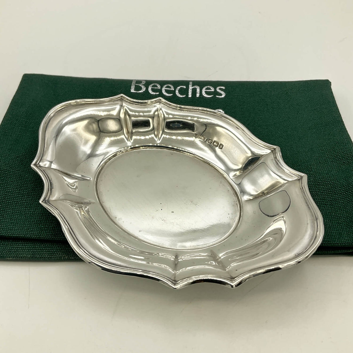 A polished silver serving dish with a scalloped rim and cutout details, displayed on a dark green Beeches Vintage cloth bag against a light background.