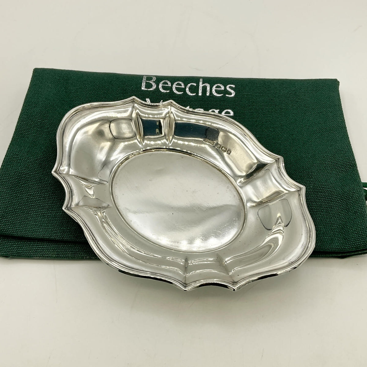 A polished silver oval plate with scalloped edges, displayed on top of a dark green Beeches Vintage cloth bag against a light background.
