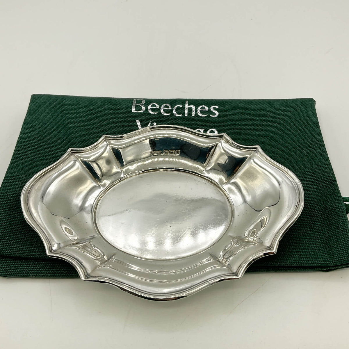 A polished silver serving tray with a scalloped rim, displayed on top of a dark green fabric pouch with the brand name 'Beeches Vintage' partially visible.