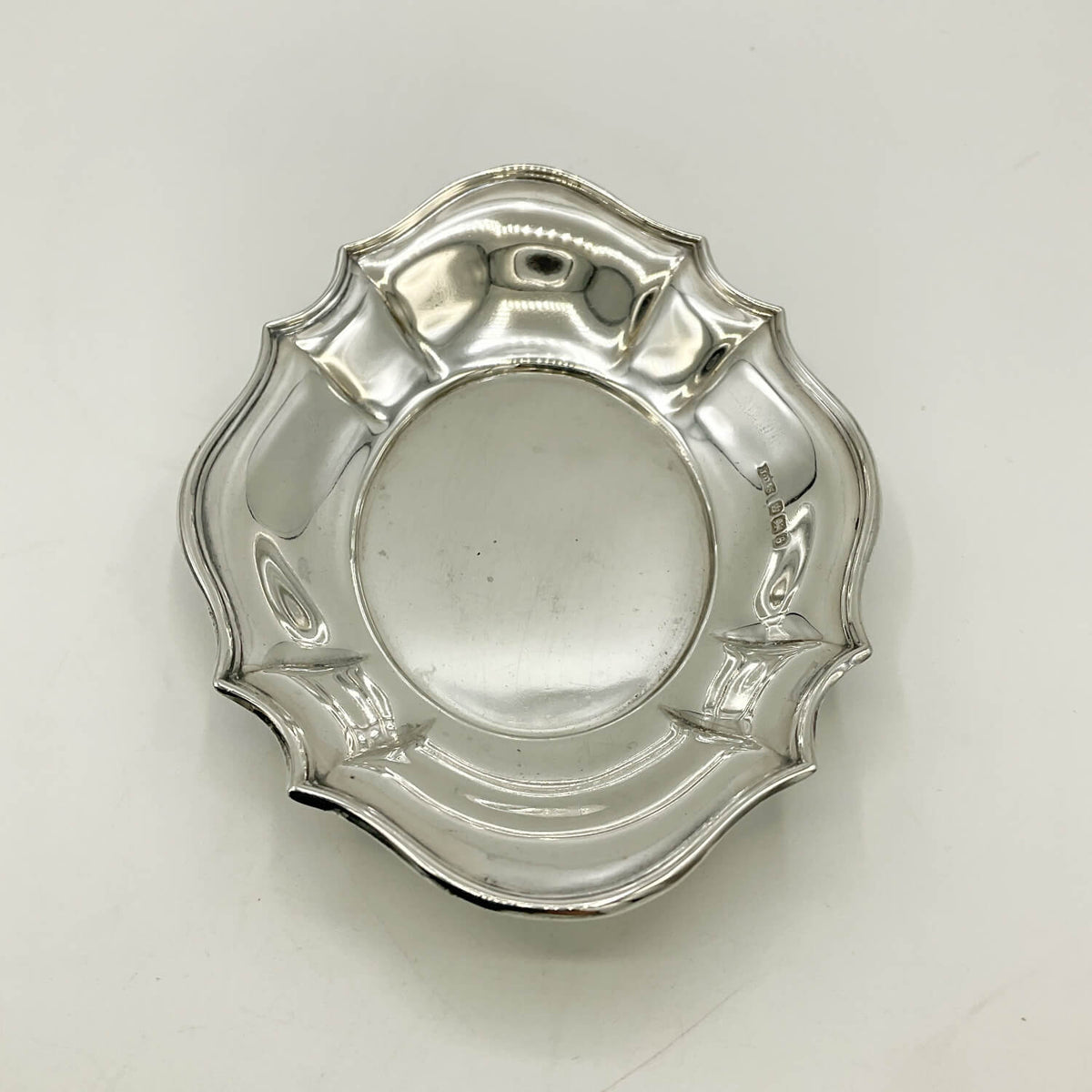 Antique sterling silver scalloped plate or dish with a reflective polished finish, photographed from directly above on a light off-white background.
