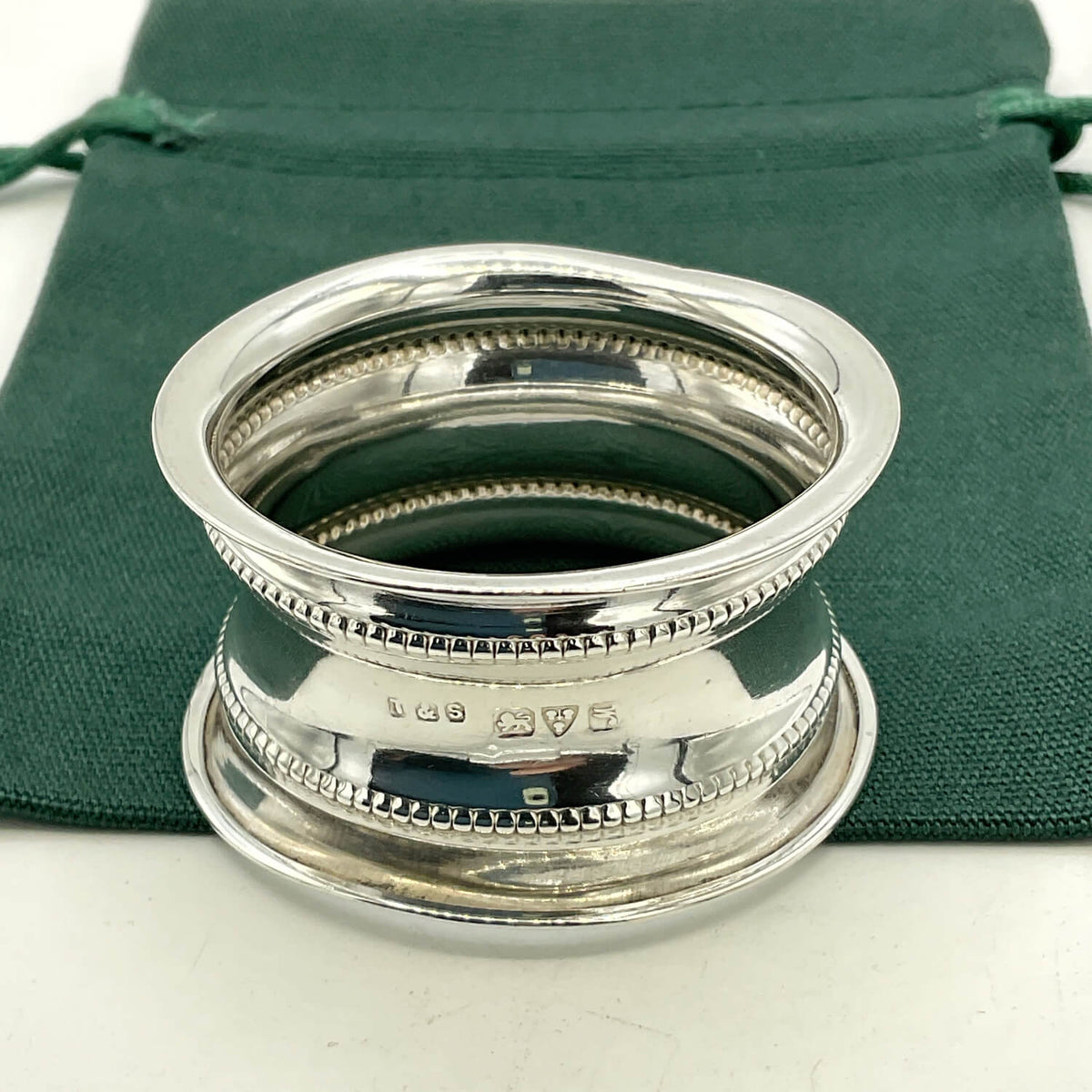 A polished silver napkin ring with decorative beaded bands, shown on a dark green velvet drawstring pouch against a white background.