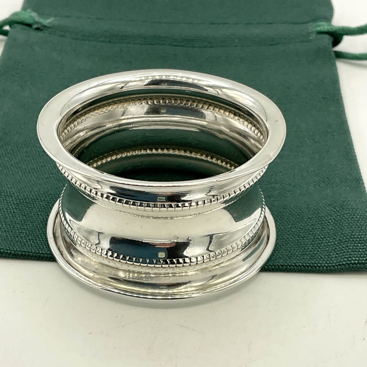 A polished silver  napkin ring with beaded decorative rims, shown resting on a dark green fabric drawstring pouch against a light neutral background.