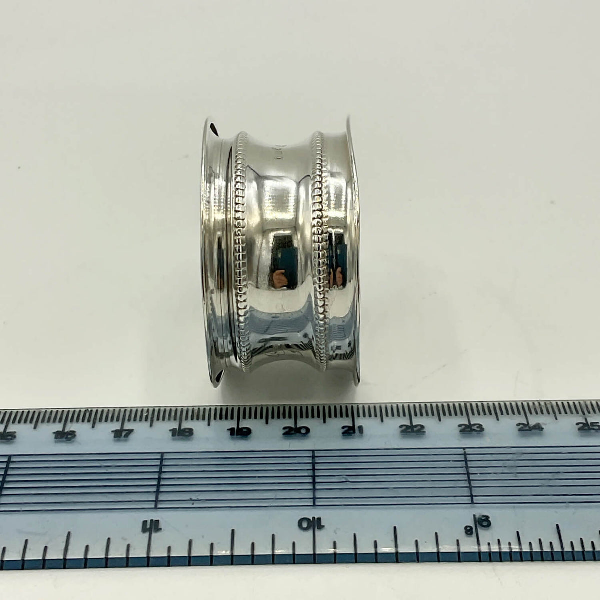 A polished silver napkin ring with decorative beaded bands, shown standing upright next to a blue metal ruler for scale on a white background.
