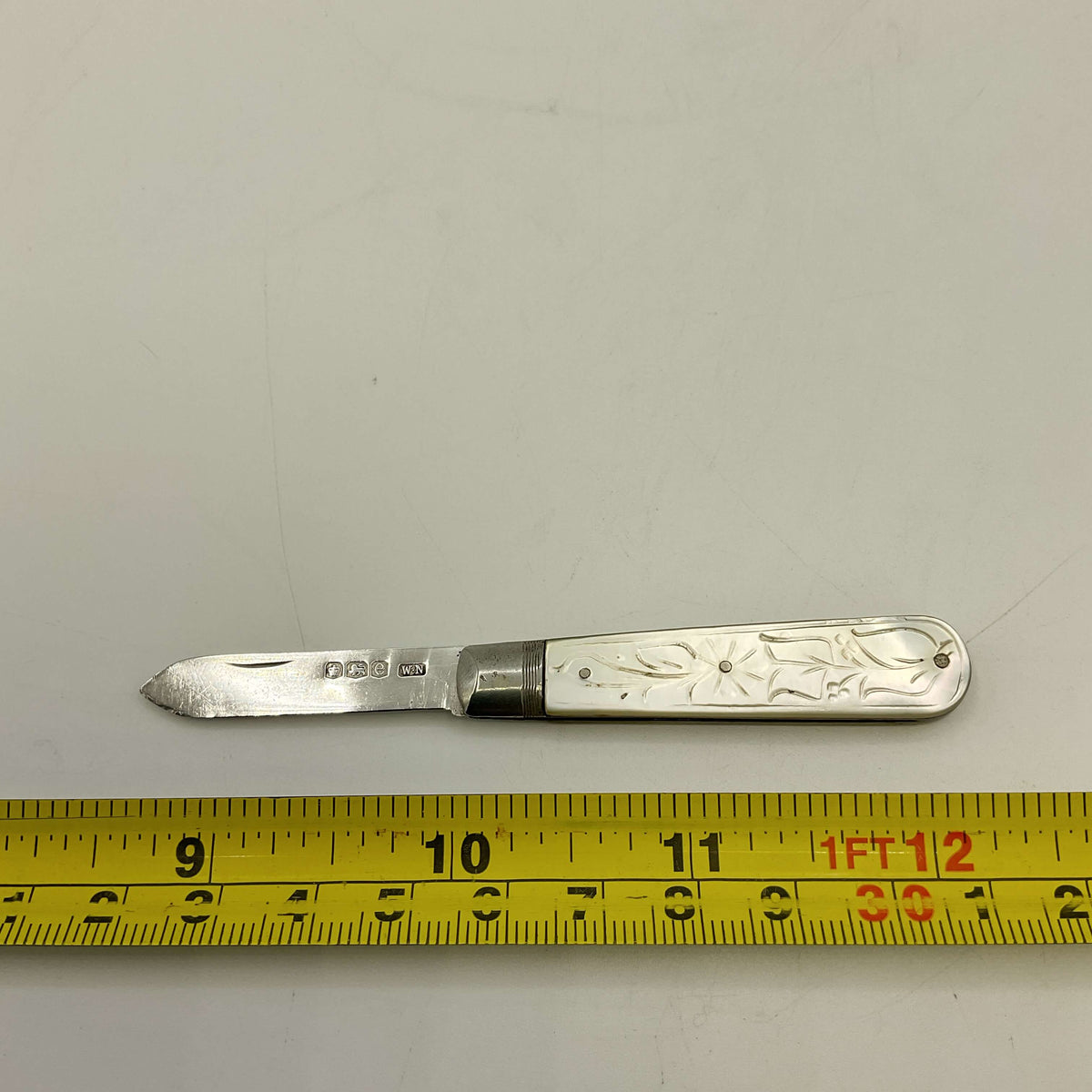 Silver fruit knife with floral design on a gray background, measured against a yellow ruler.