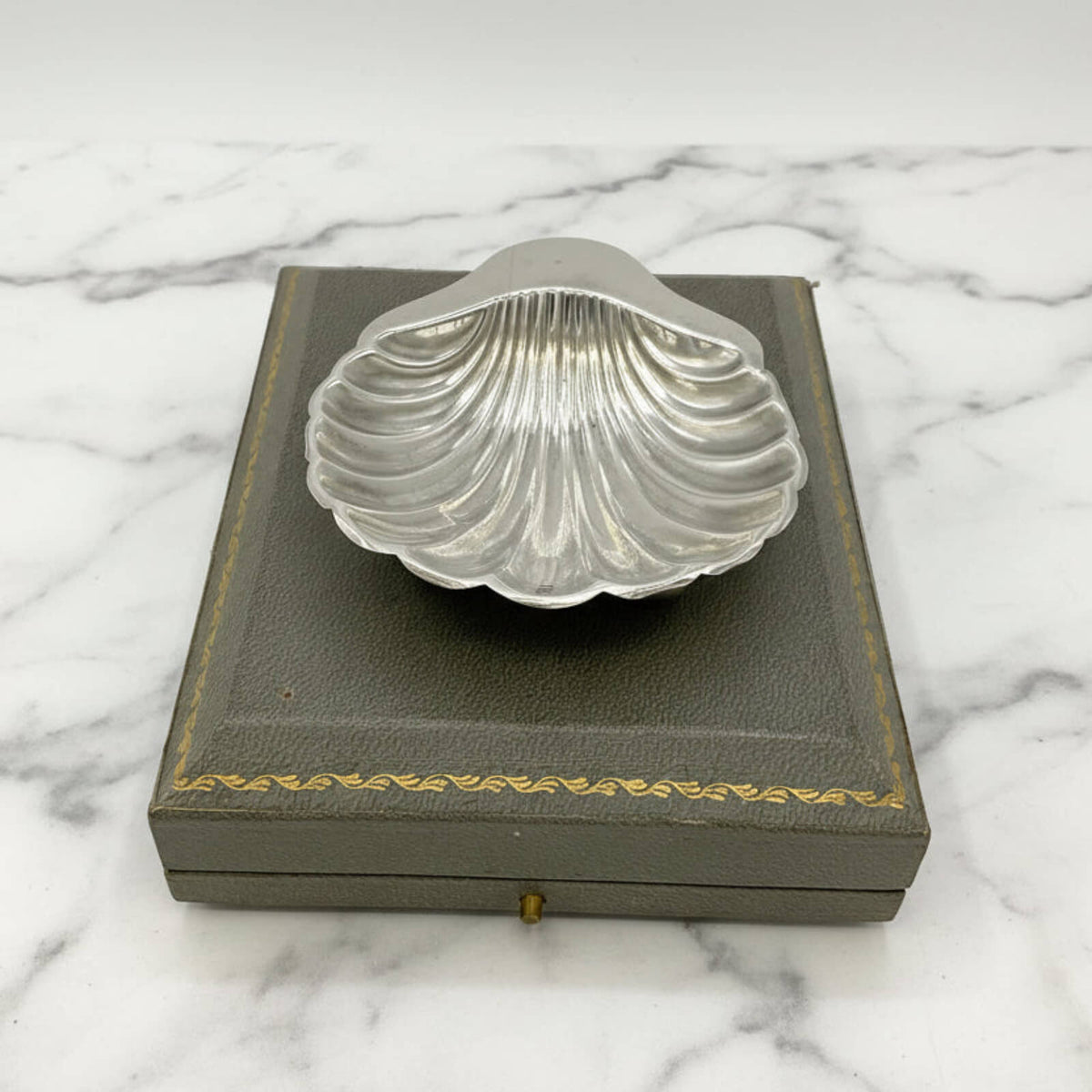 Silver shell-shaped dish on a gray box with gold trim against a marble background