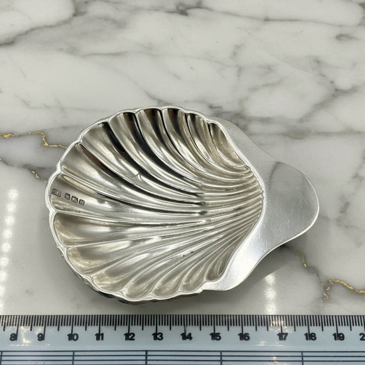 Silver shell-shaped dish on a marble surface with a ruler for scale.