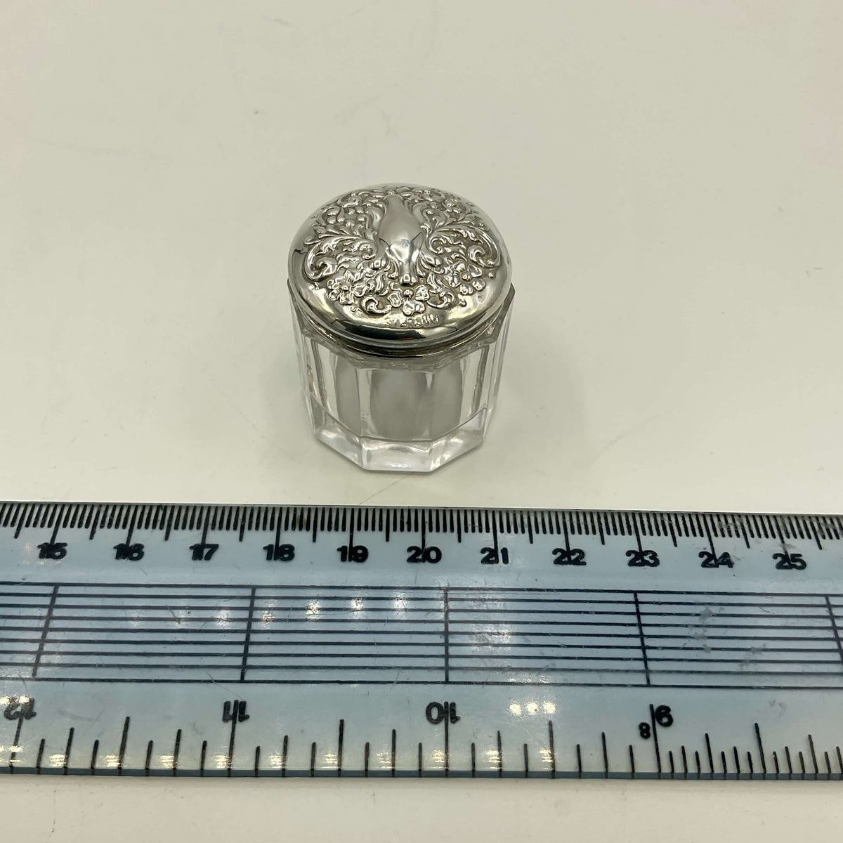 Small glass jar with silver lid on a white background with a ruler for scale.