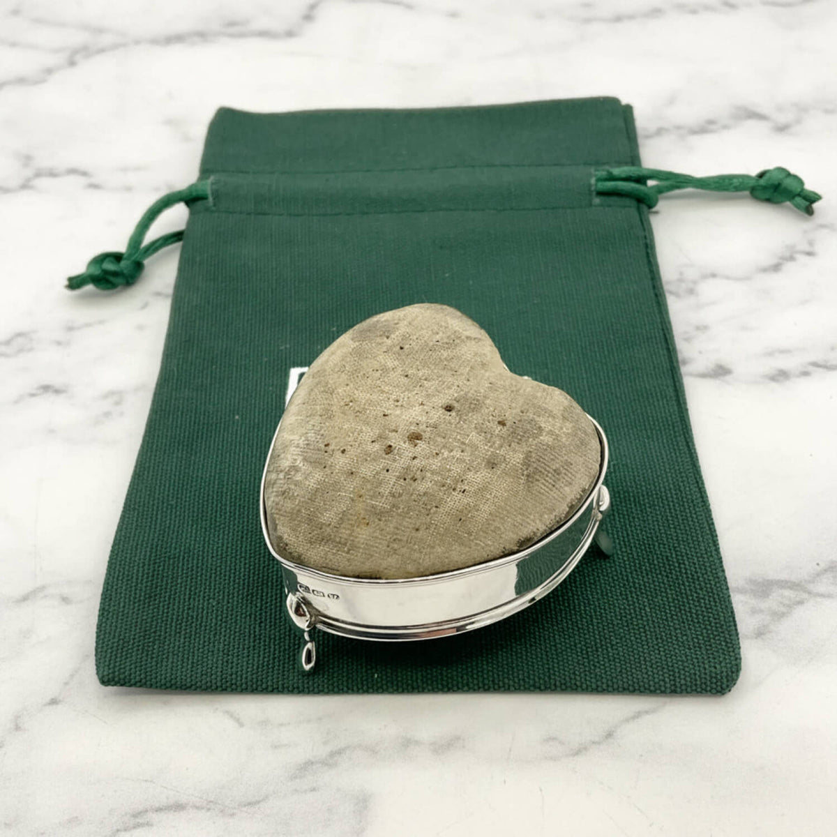 Heart-shaped pincushion in a silver holder on a green drawstring bag with a marble background
