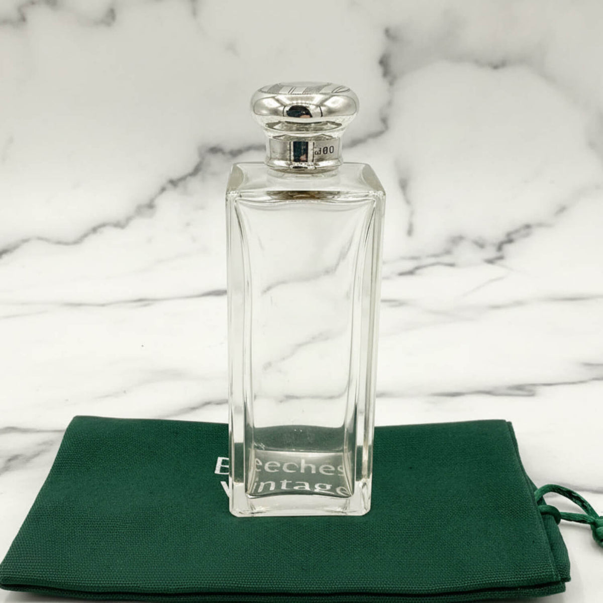 Clear glass perfume bottle with silver cap on a green cloth with 'Beeches Vintage' branding against a marble background.