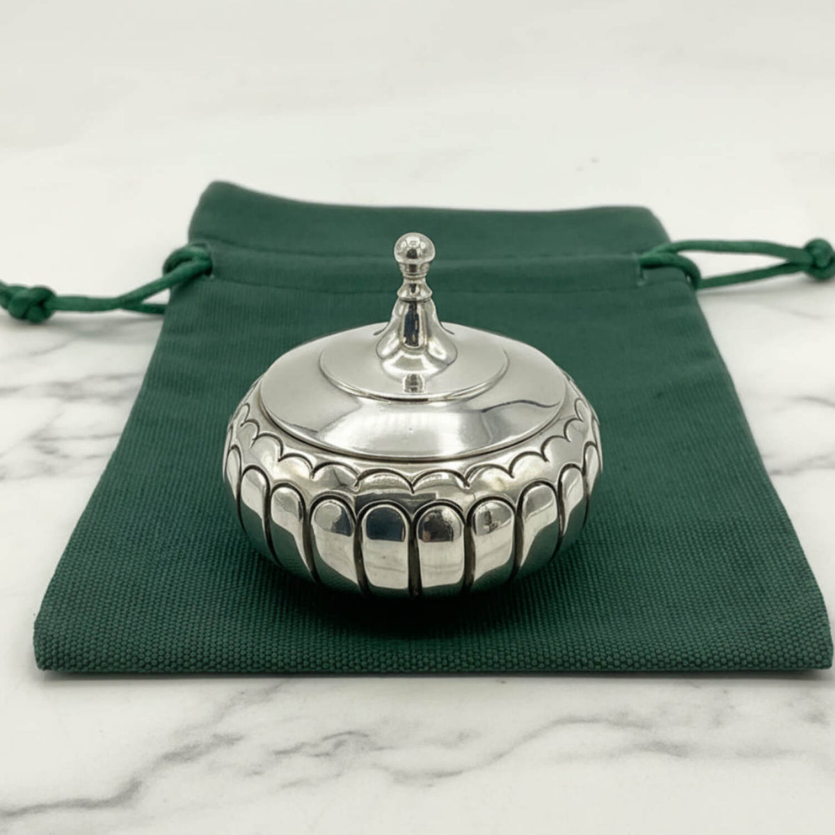 A small round silver box with a fitted lid and decorative scalloped edge, placed on top of a dark green drawstring pouch over a light marble surface.