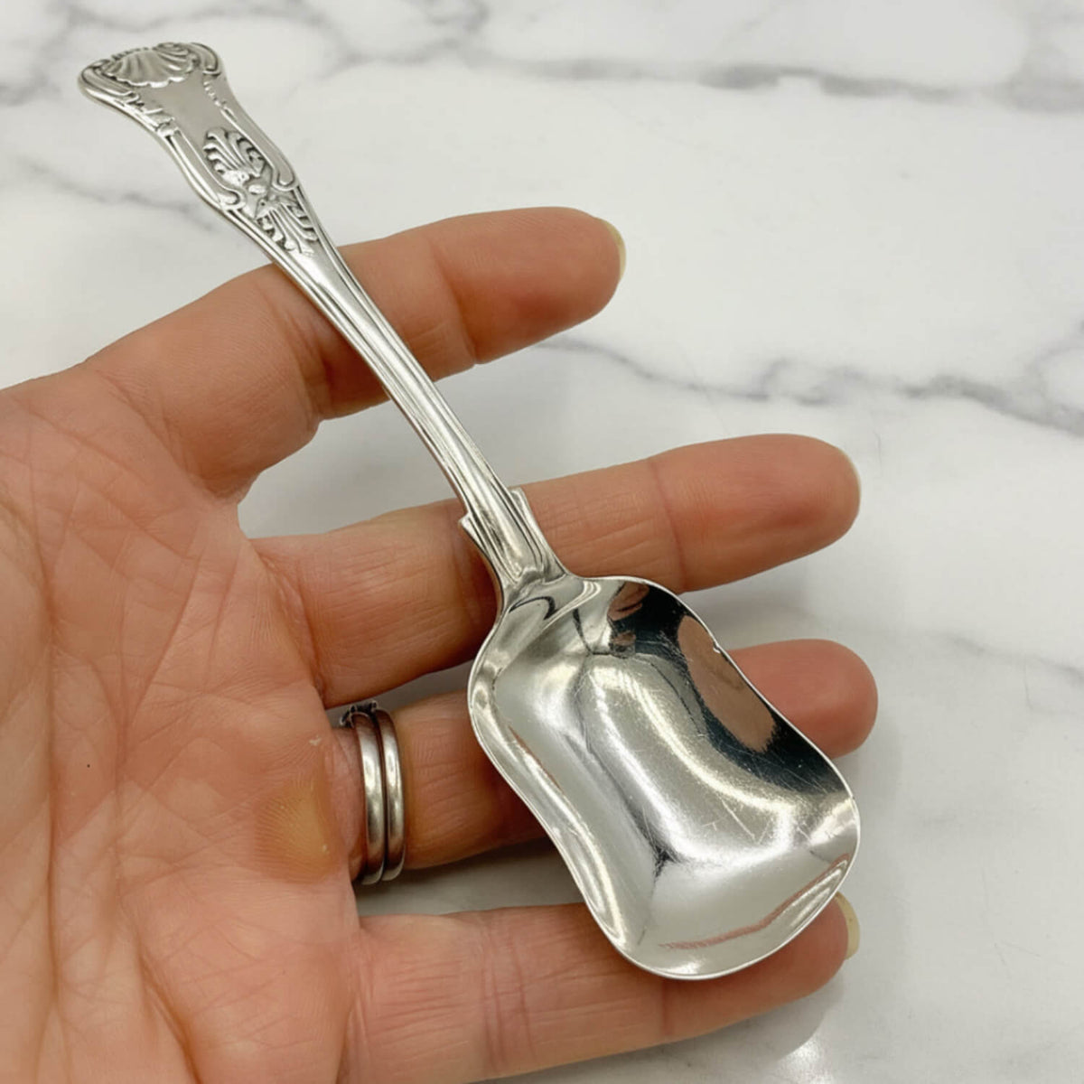 Antique Victorian Sterling Silver Spoon with an ornate handle, held in a person's hand over a white marble surface.