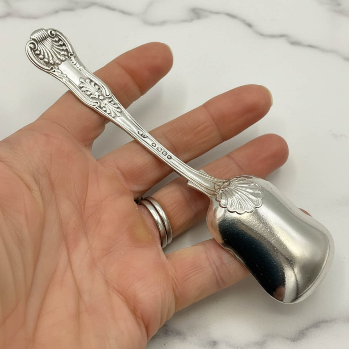 Antique Victorian Sterling Silver Spoon with an ornate engraved handle, held in a person's hand against a white marble background.