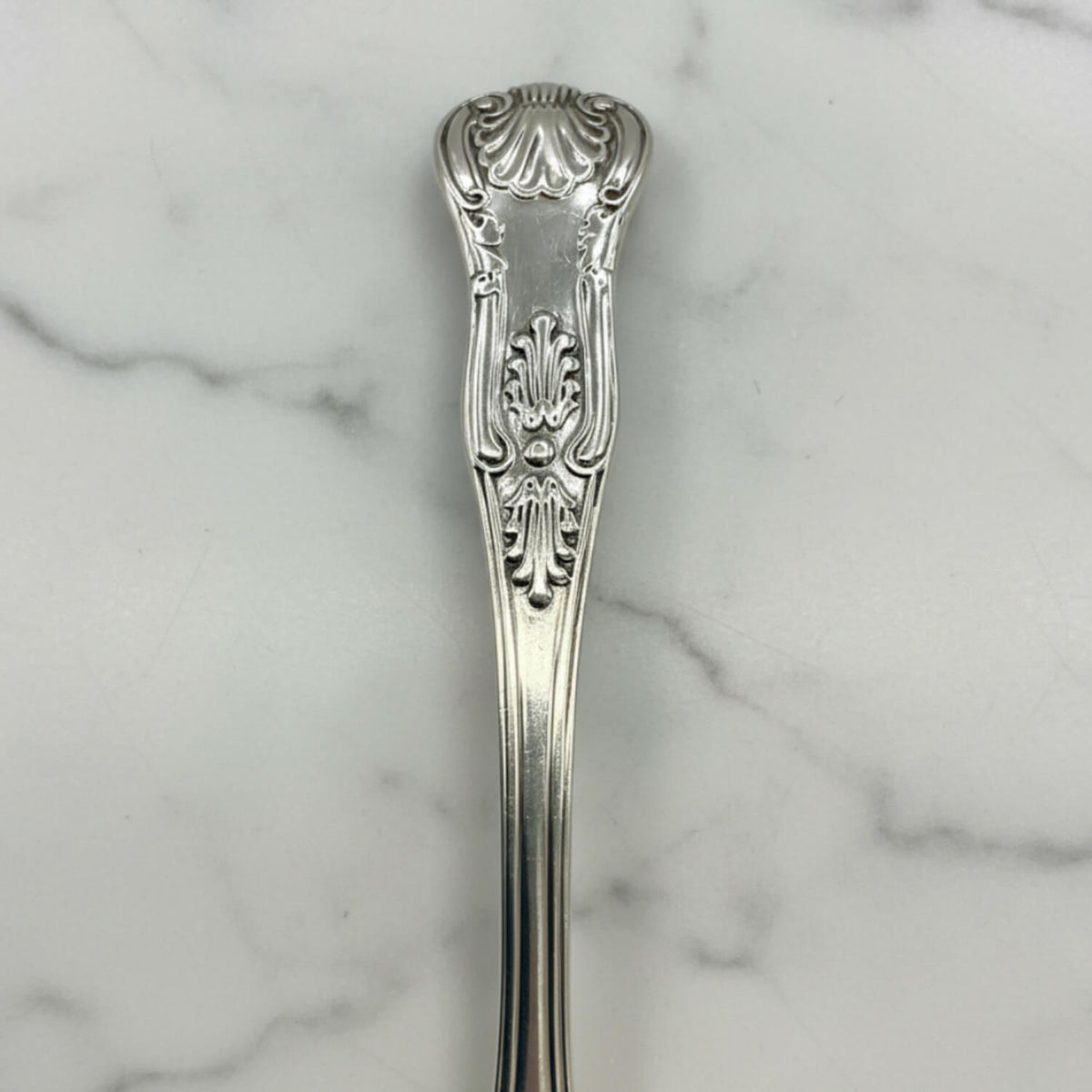 An ornate silver spoon handle, featuring a detailed floral and scrollwork pattern, resting on a white marble surface.