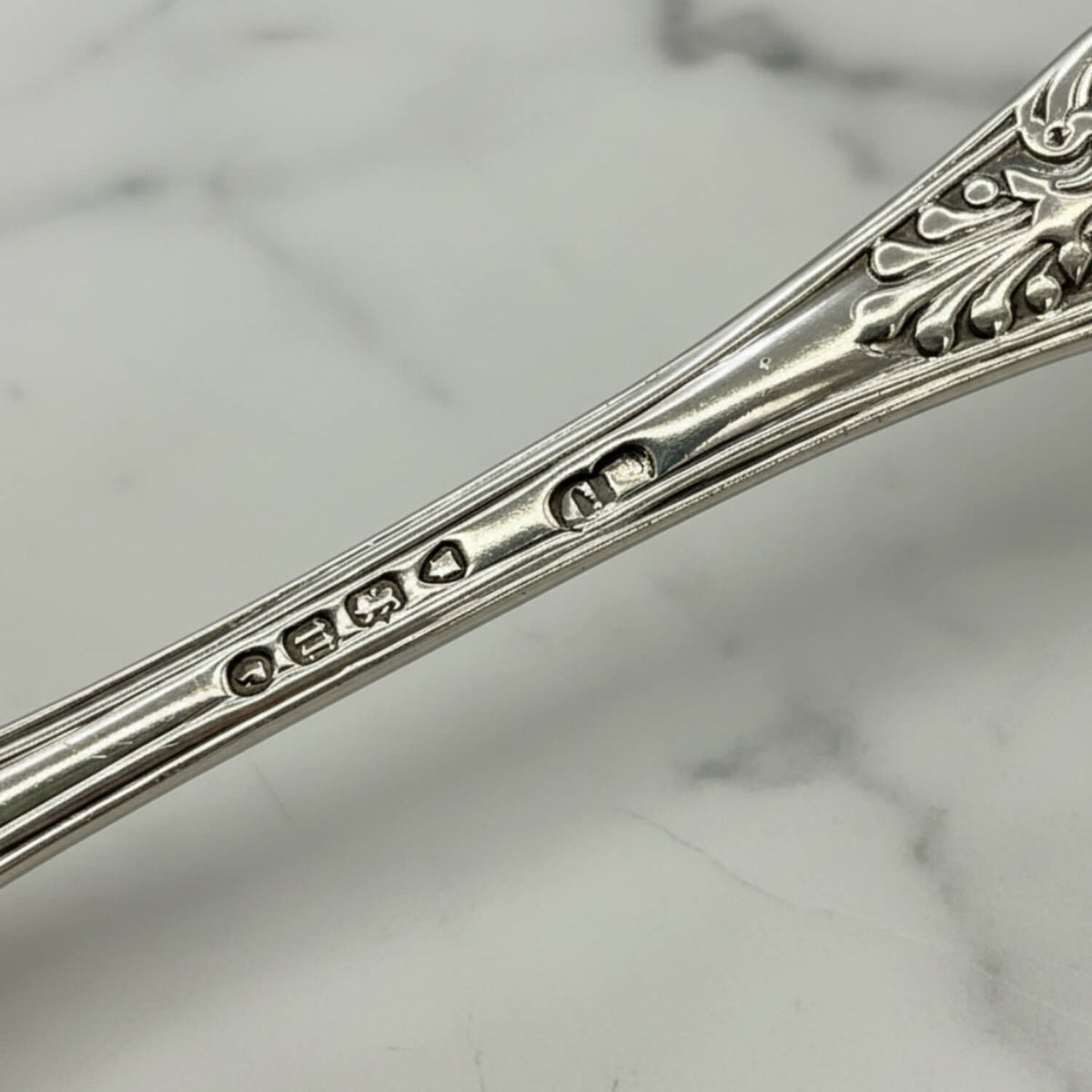 Close-up of Antique Victorian Sterling Silver Spoon handle with engraved decorative detailing and maker's marks, resting on a white marble surface.