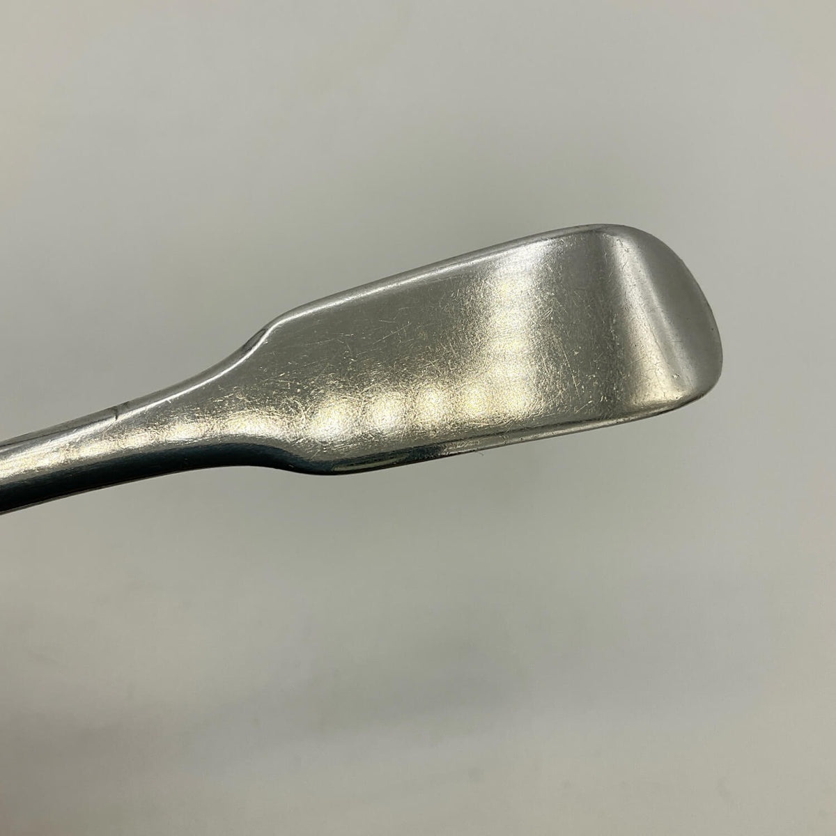 A close-up of a silver teaspoon or spatula head on a plain light gray background.