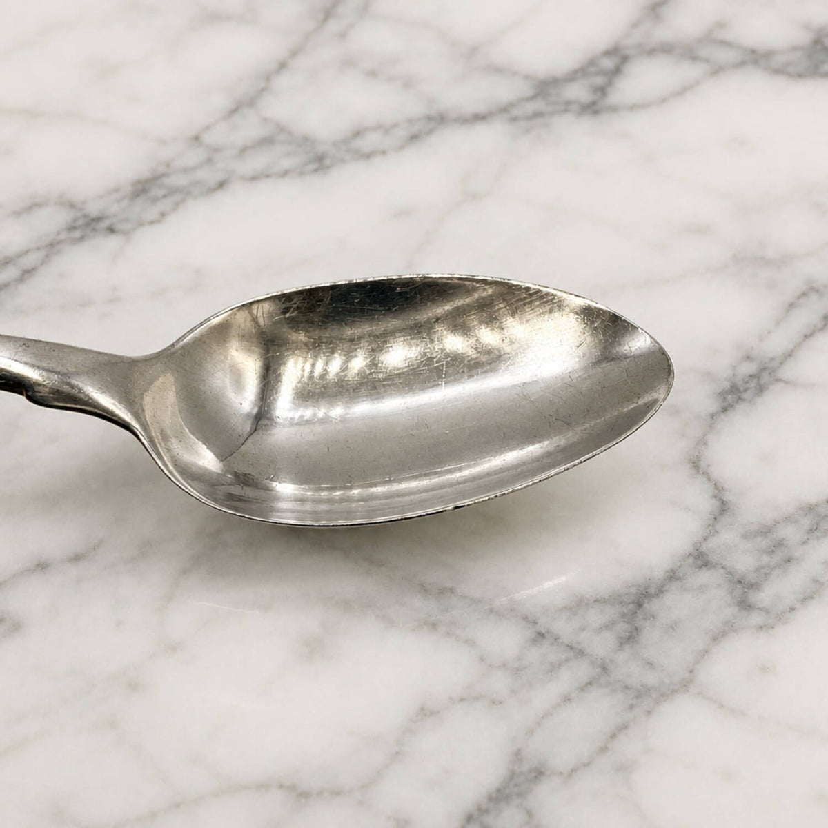 A polished silver teaspoon shown in profile against a marble light gray background.