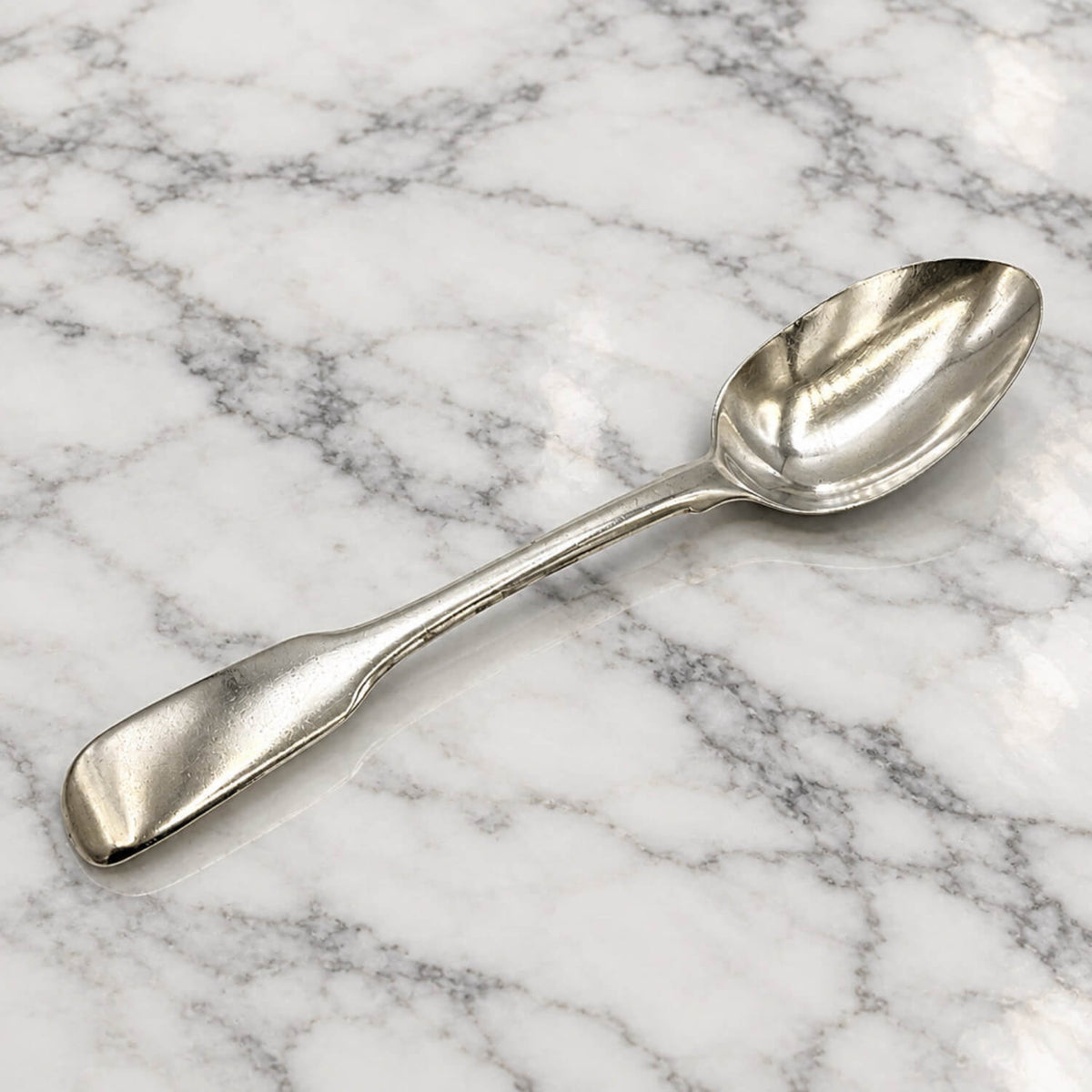 A polished silver teaspoon resting diagonally on a white and gray veined marble surface.