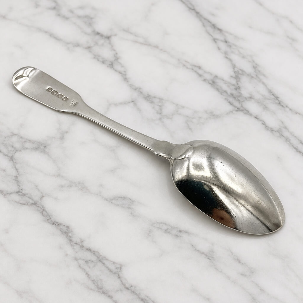 An antique sterling silver teaspoon with a polished finish, resting diagonally on a white marble surface with gray veining.