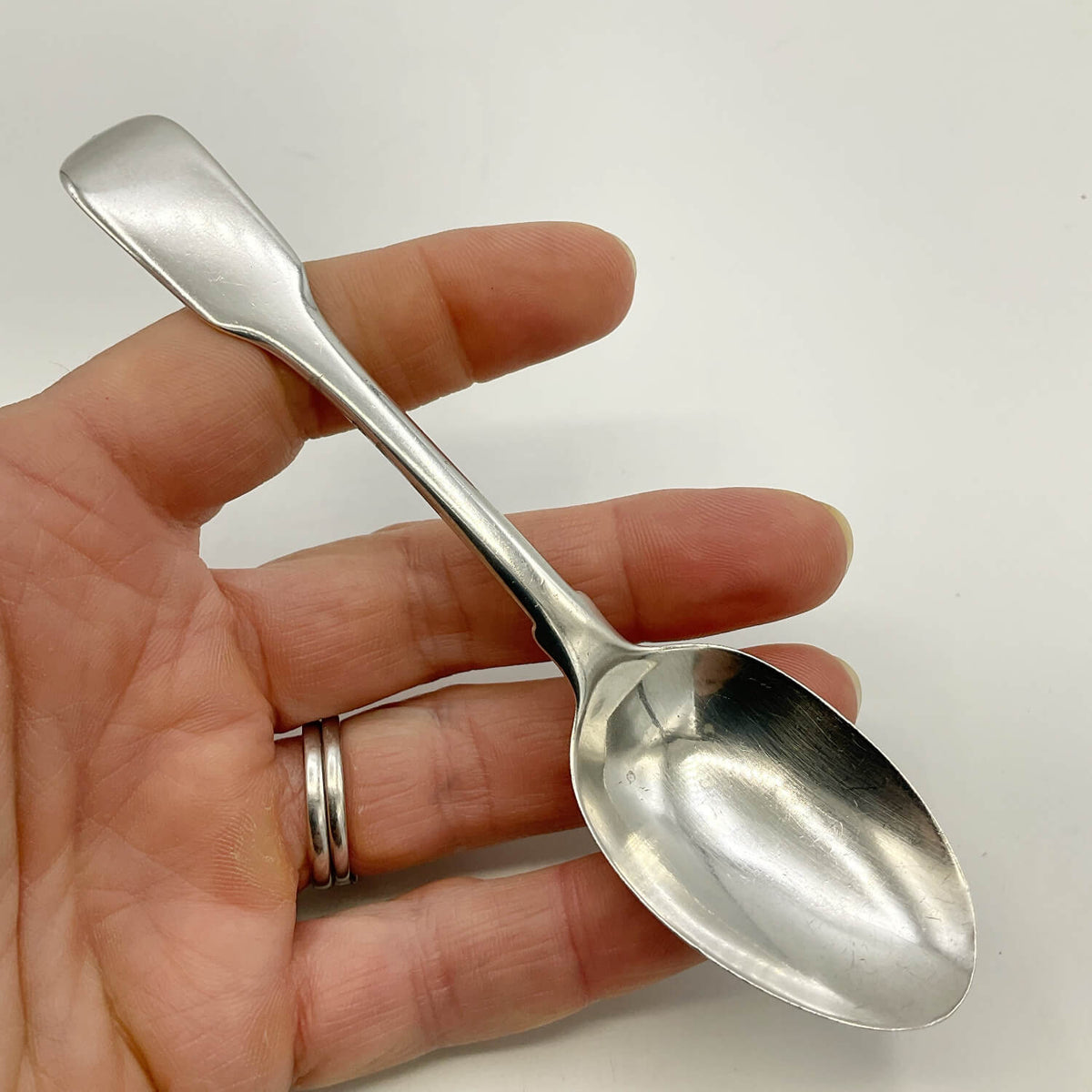 A polished silver teaspoon with a flat, rectangular handle, held in a person's hand against a plain white background.