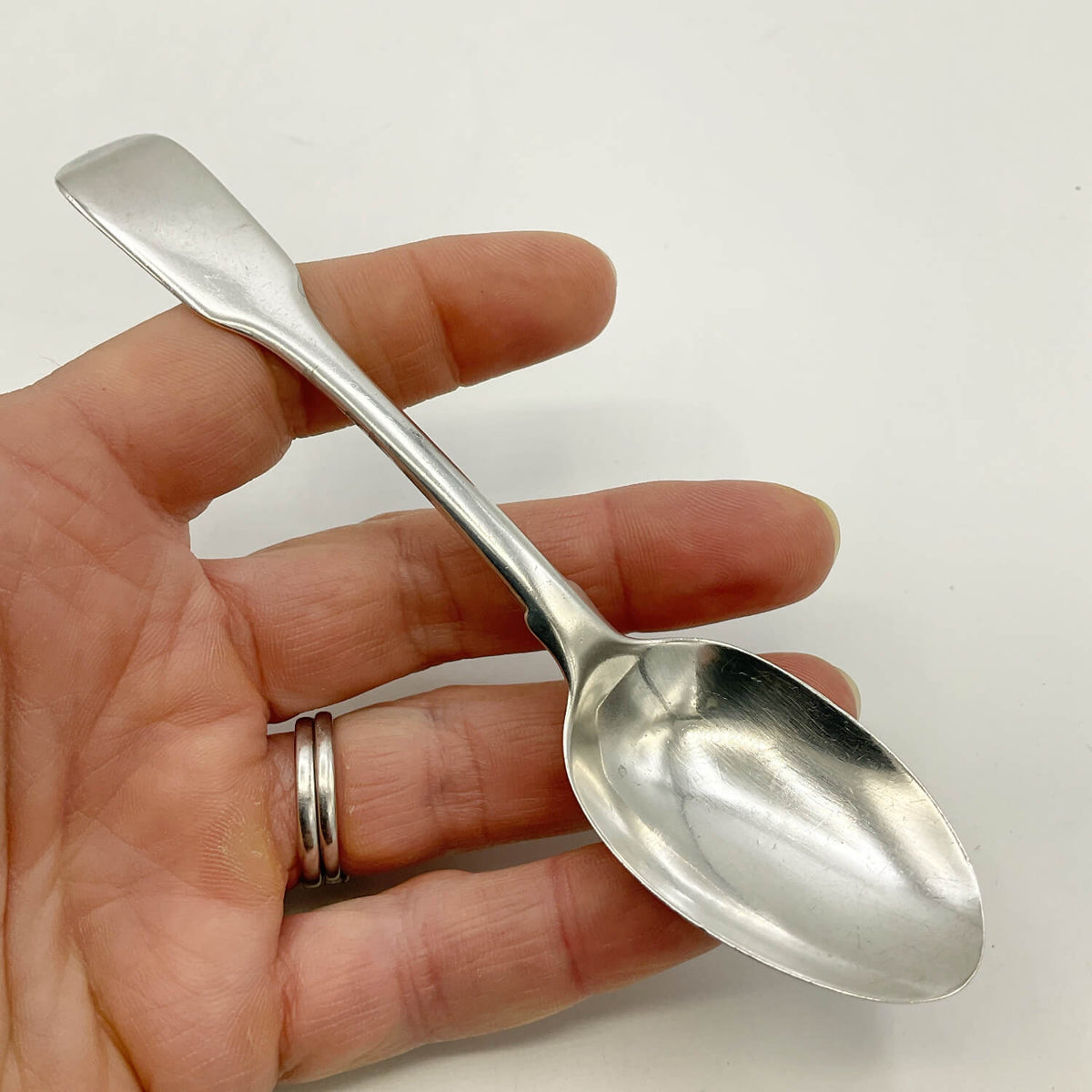 A polished silver teaspoon held in a person's hand against a plain white background.