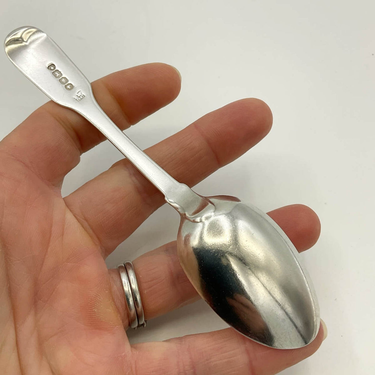 A polished silver spoon held in a person's hand against a plain white background. The spoon has a small engraved maker's mark and hallmarks on the handle.