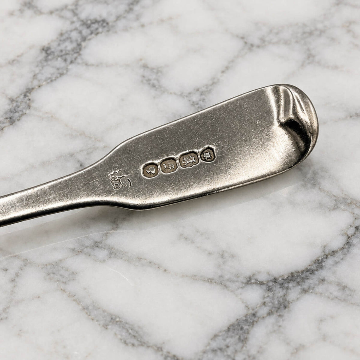 A close-up of a silver teaspoon handle resting on a white and gray marble surface, showing engraved hallmarks on the back.
