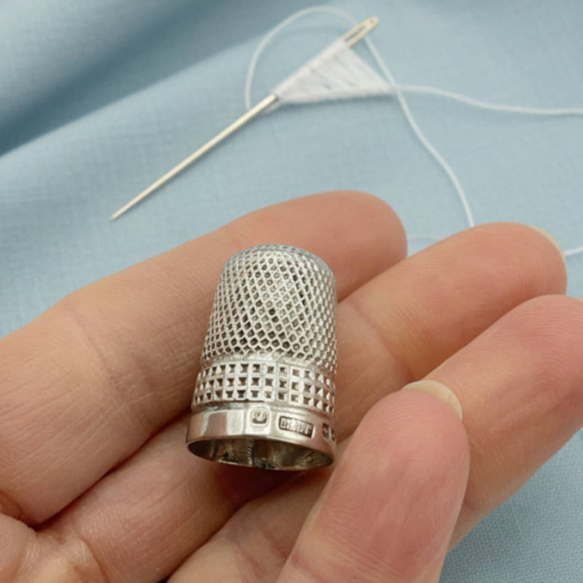 Silver thimble held in a hand with a needle and thread in the background