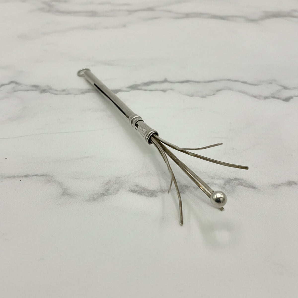 A silver Art Deco Sterling Silver Cocktail Whisk with a textured handle, a small ball tip, and several thin prong-like wires, photographed on a white marble surface.
