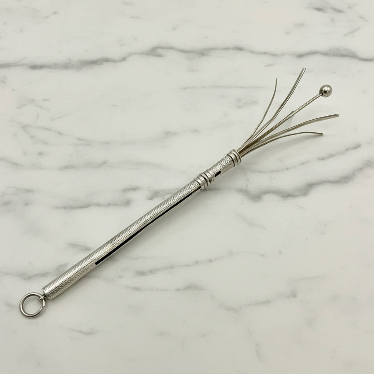 A silver metal cocktail whisk with a textured handle, a hanging loop, and fine wire bristles ending in a small ball, photographed on a white marble surface.
