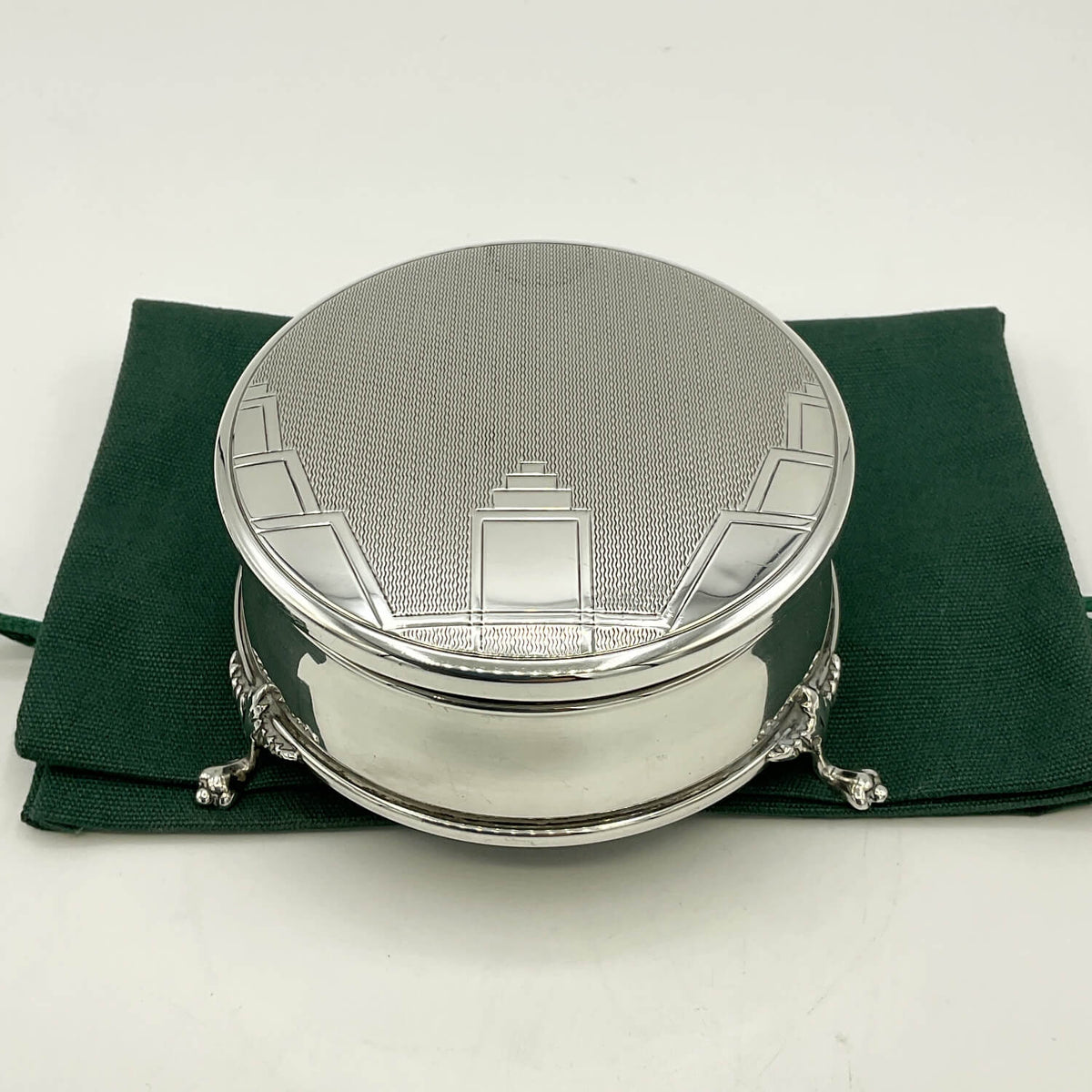 A round silver box with a textured lid and decorative embossed Art Deco details, resting on a dark green fabric pouch against a plain white background.