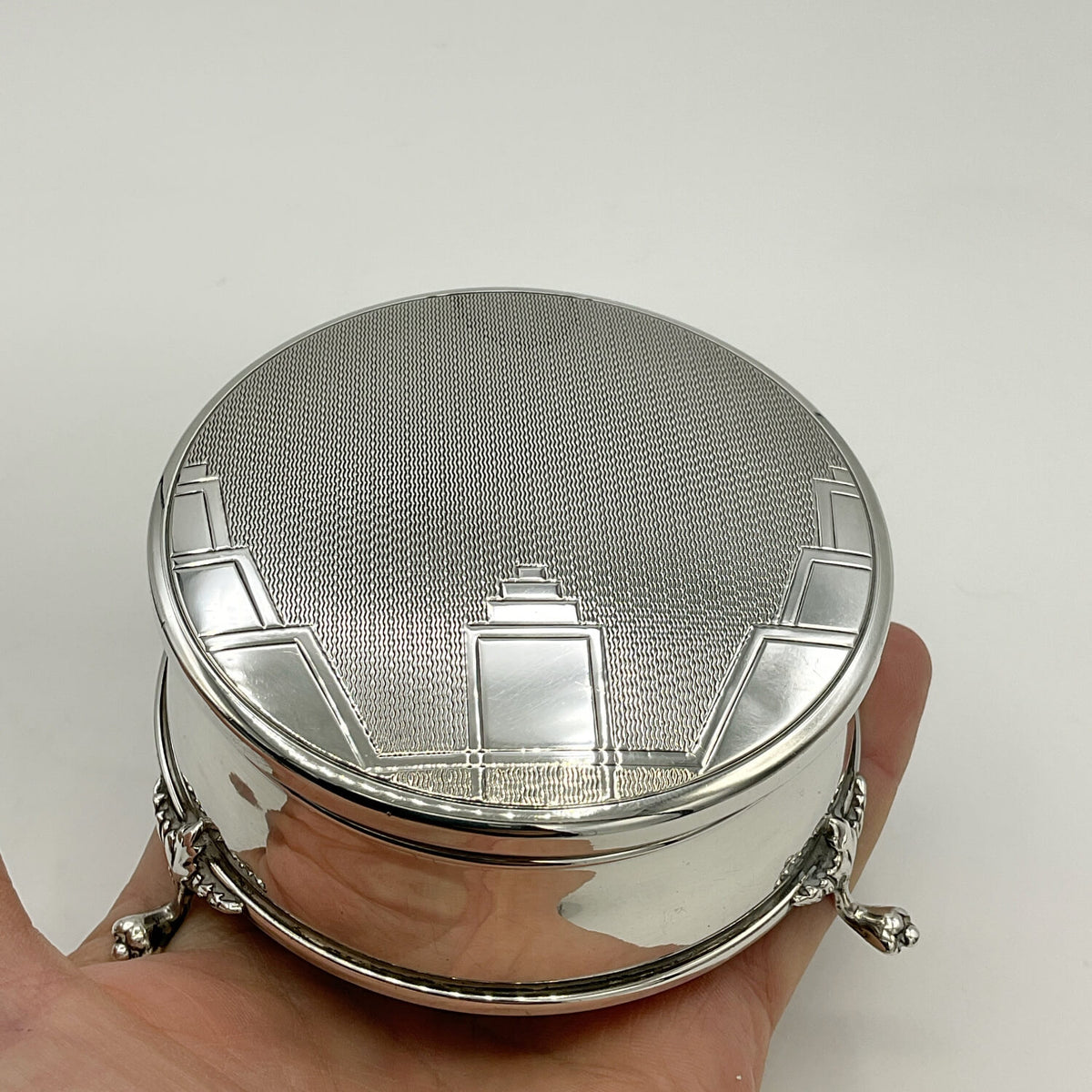 A round silver box with a textured lid and decorative geometric engravings, shown resting on a person's hand against a plain light background.