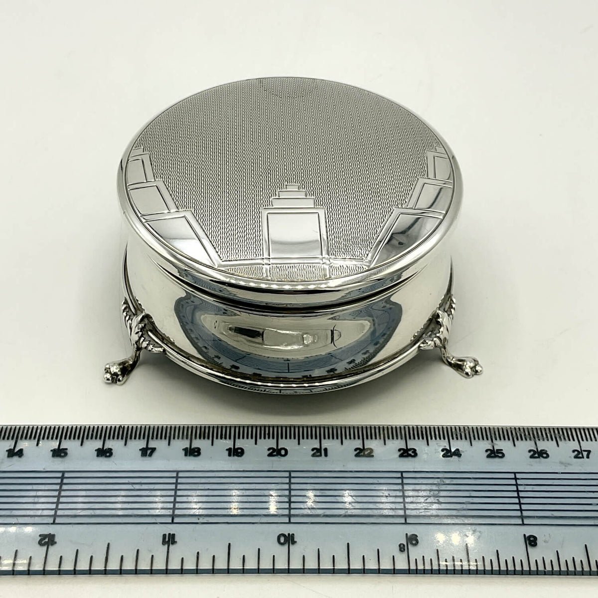 A small round silver box with a textured lid and decorative architectural engraving, shown on a white background with a ruler at the bottom for scale.
