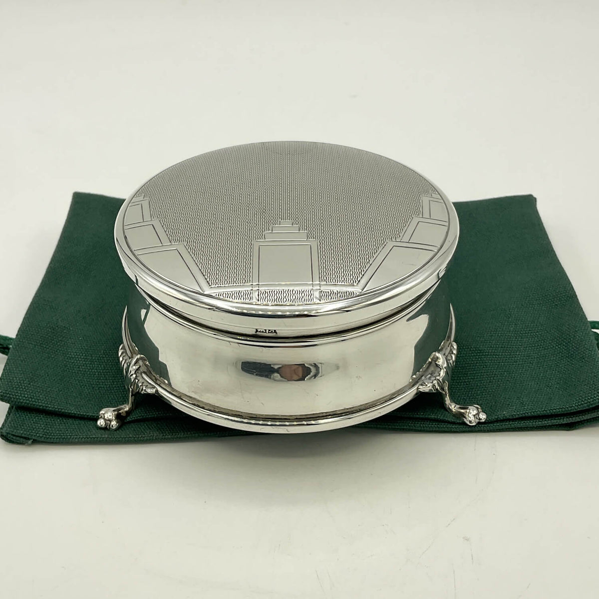 A round silver lidded box with ornate claw-style feet and a textured lid featuring an engraved building design, displayed on a dark green fabric pouch against a white background.