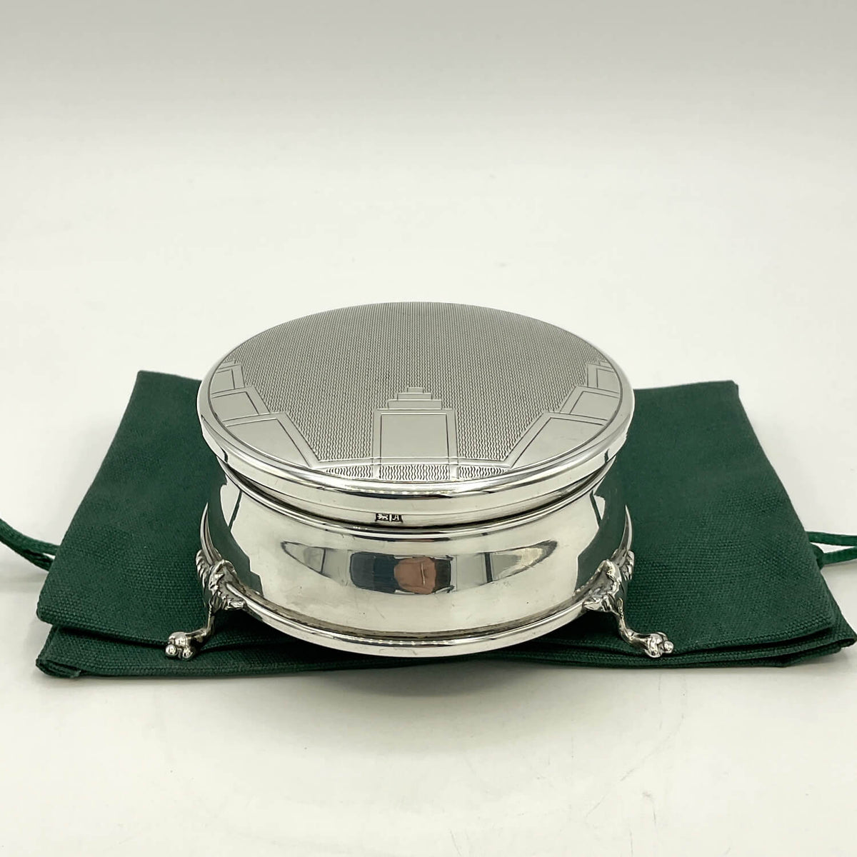 A round silver box with a textured lid and small feet, displayed on a dark green velvet pouch against a white background.