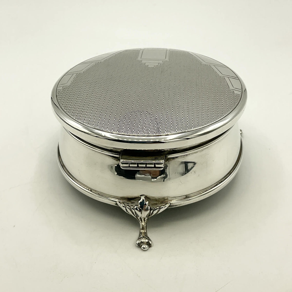 A polished silver round case with a textured, patterned lid and a decorative three-pronged stand, shown on a plain white background.