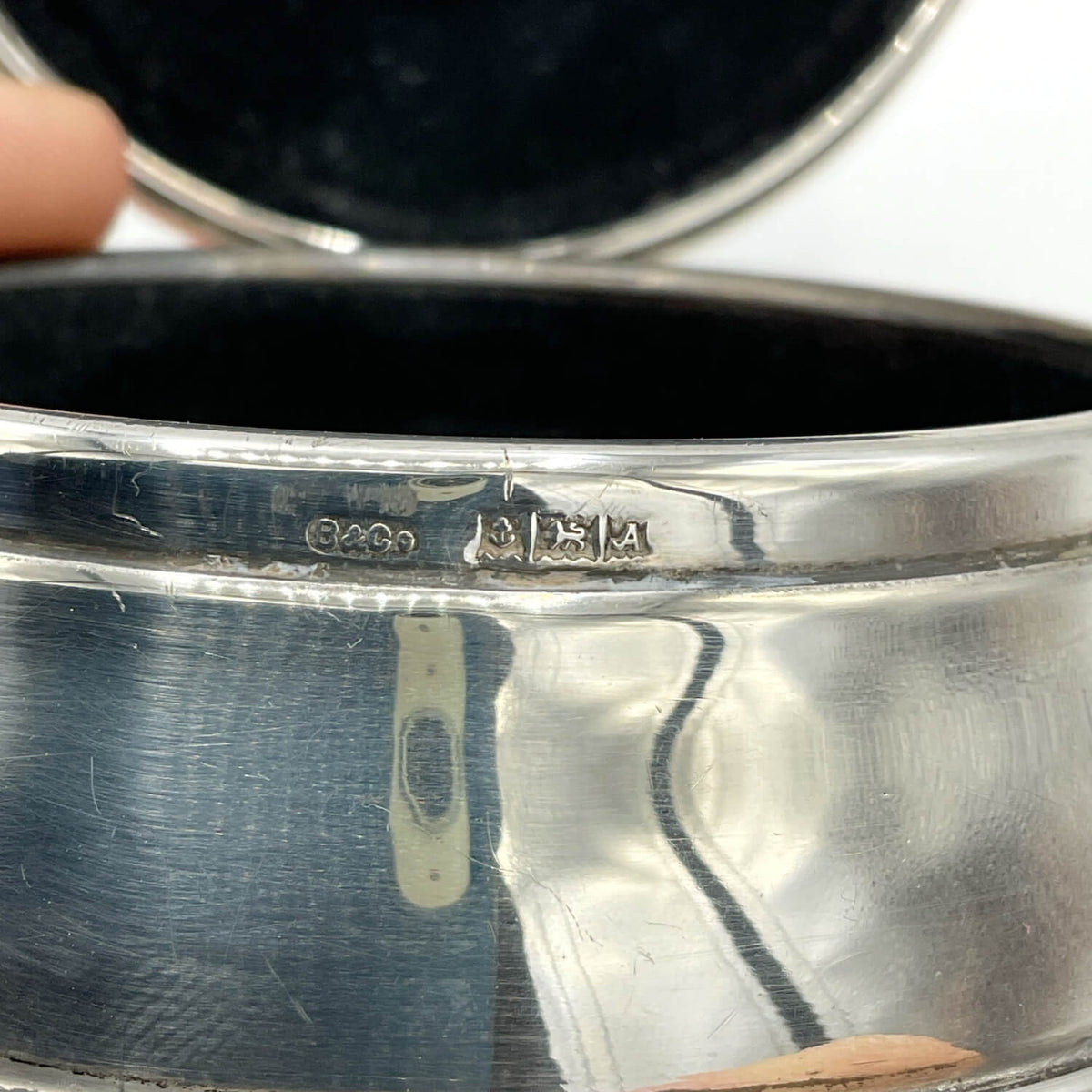 A close-up of a silver jewellery box held by fingers, showing the inner band with engraved hallmarks and a dark velvet-lined interior.